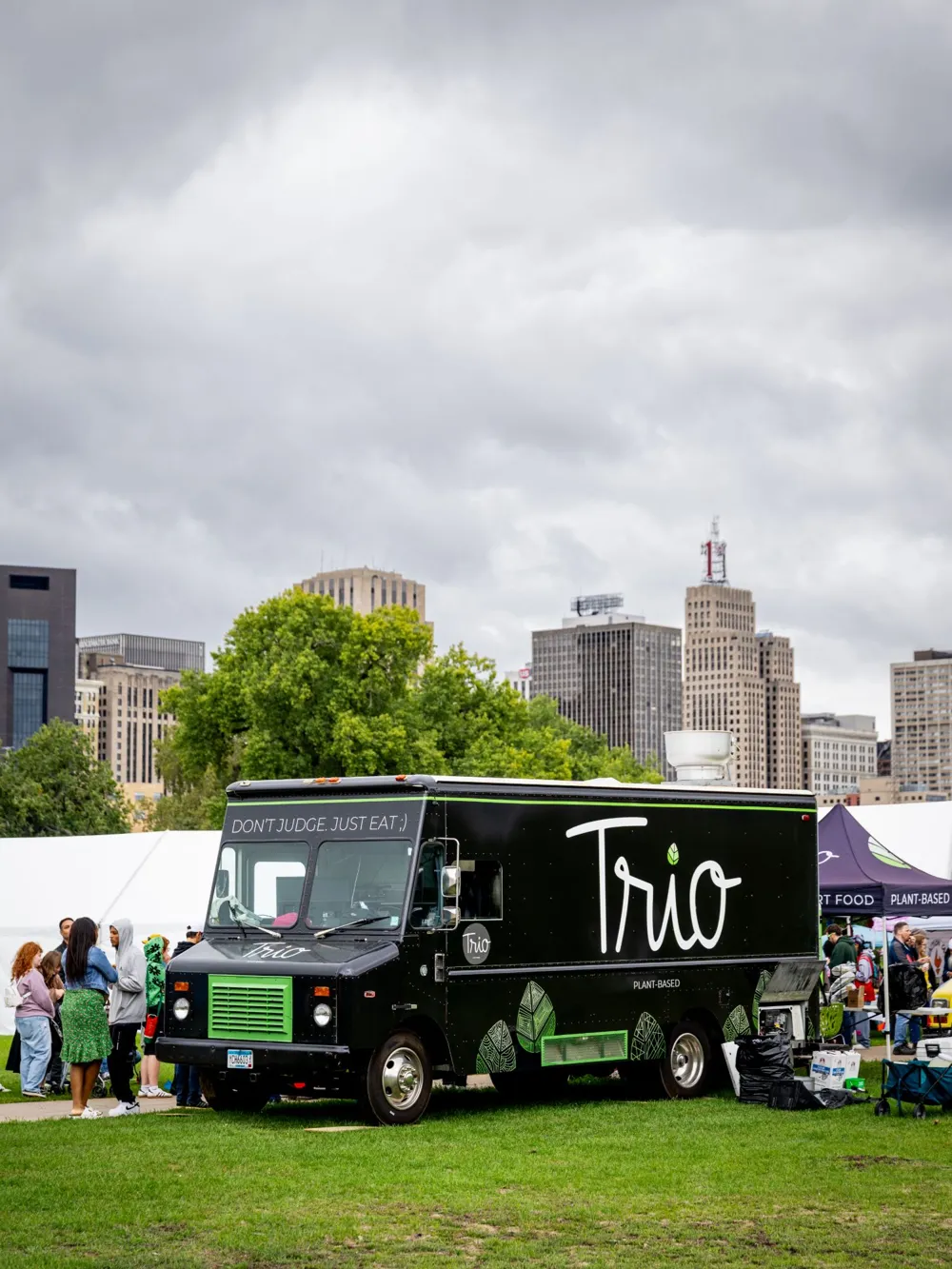 The black Trio Plant-Based food truck parked on the grass at an outdoor festival, with the St. Paul skyline behind it